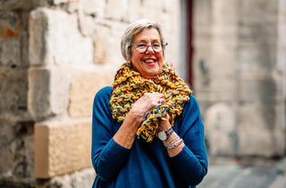 Woman wearing beautiful yellow, blue and red hand knitted woollen scarf and blue merino jump from the Salamanca Wool Shop. The woman is standing in front of a sandstone wall in Salamanca, Tasmania.